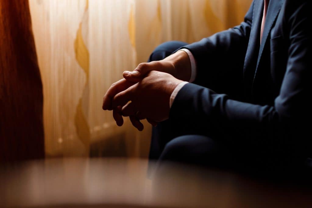 An adult in a dark suit sits with their hands clasped in front of a window
